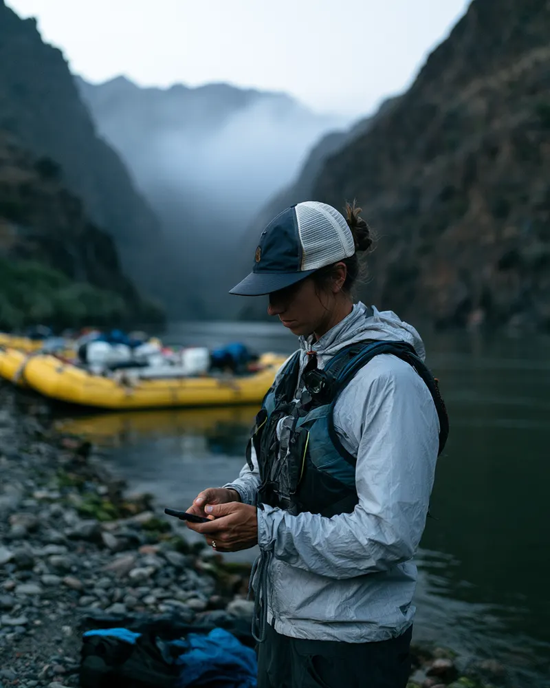 Rafting guide checking a phone beside a misty river at dawn
